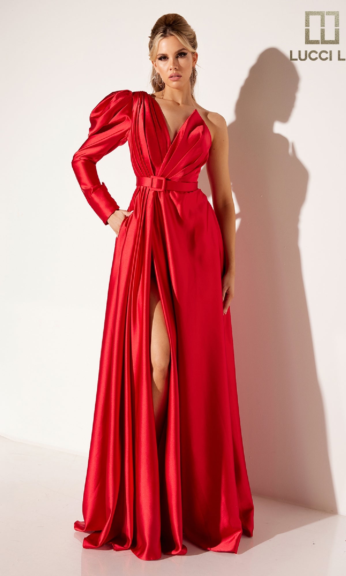 Wearing the Lucci Lu C8098 long formal dress, a woman stands by a white wall in an elegant red gown featuring a v-neckline, thigh-high slit, and one long puffed sleeve.